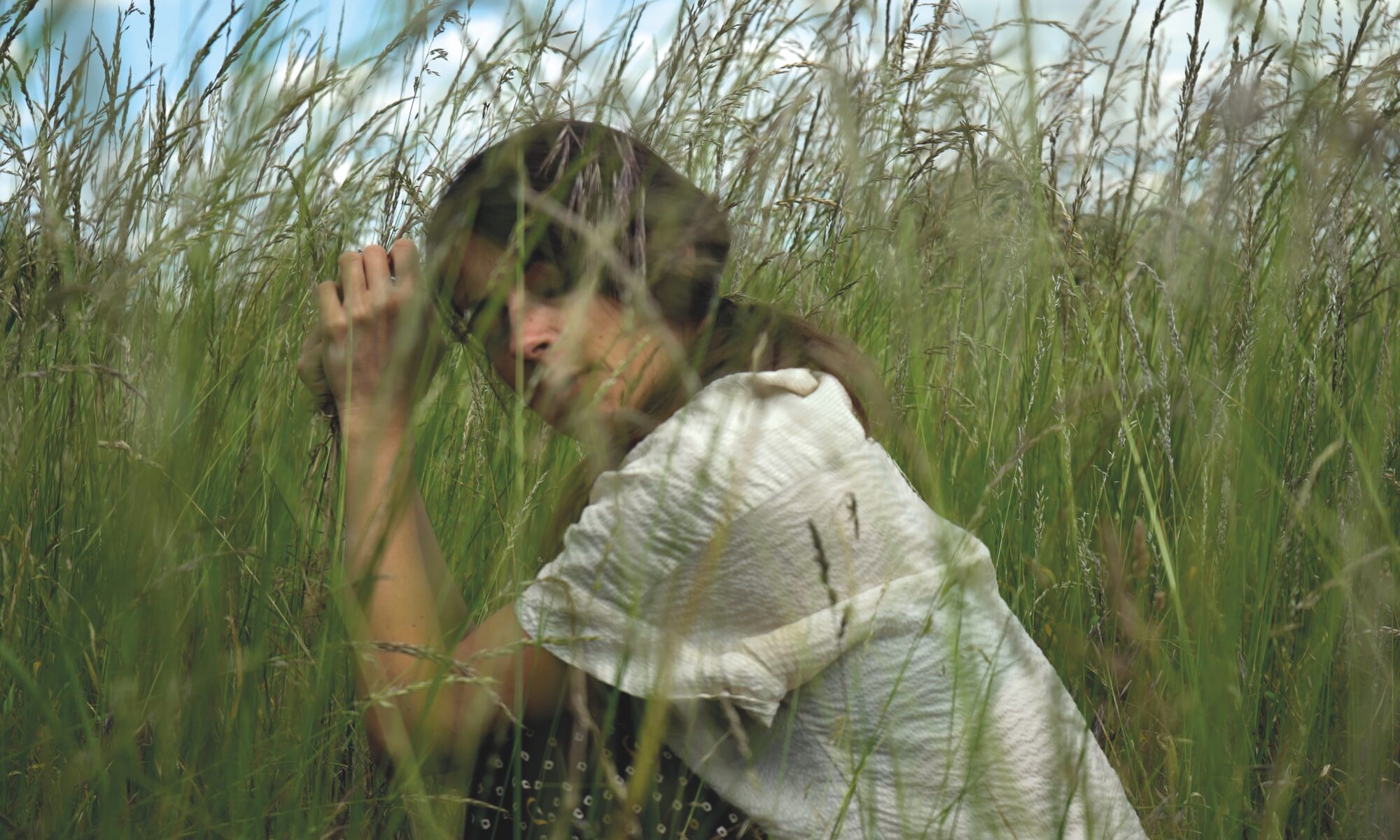 Claire is crouching eyes closed in the middle of very tall grasses which are taller than her in this position. She looks peaceful and immersed, totally in tune with natural world that she is part of.
