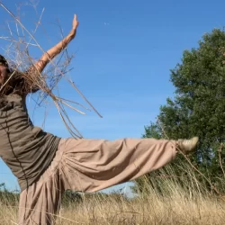 Claire is making a star shape with her right leg standing on the ground and the left at a perpendicular position off the ground. Her mouth is wide open and one of her harm carries dried cow parsley all other. She's standing in the middle of Hackney Marshes at the end of the summer when the grass has turned yellow and dry.